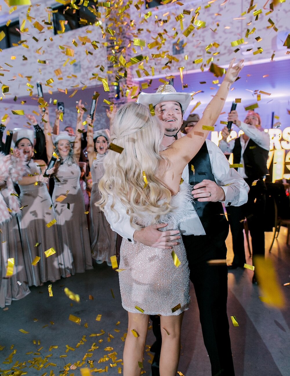 The bride and groom hug and dance under a shower of falling gold confetti, with the groom wearing a white cowboy hat and vest during the reception.