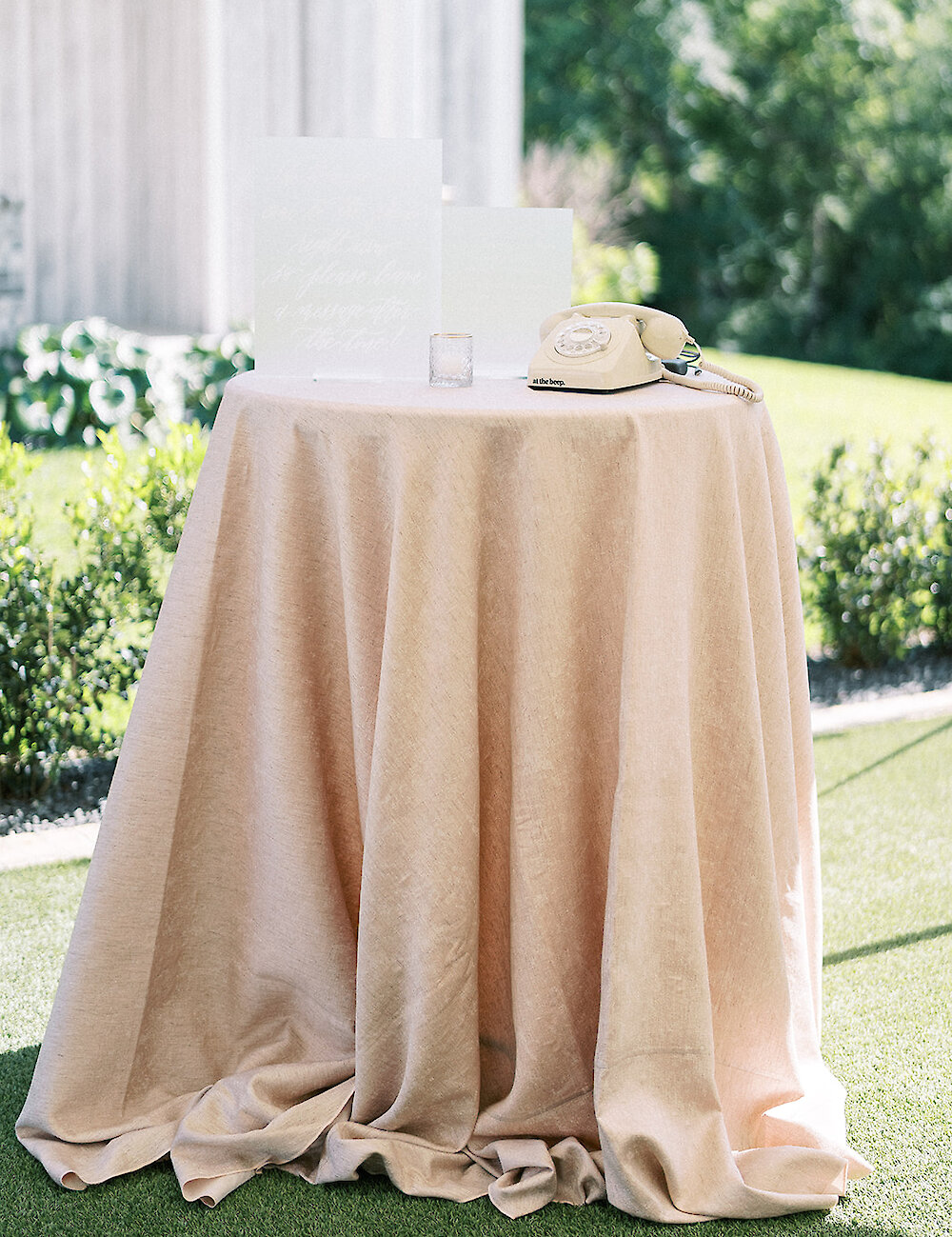 A vintage cream-colored telephone sits on a small round table covered with a blush linen tablecloth for use as an audio guest book outdoors on the lawn.