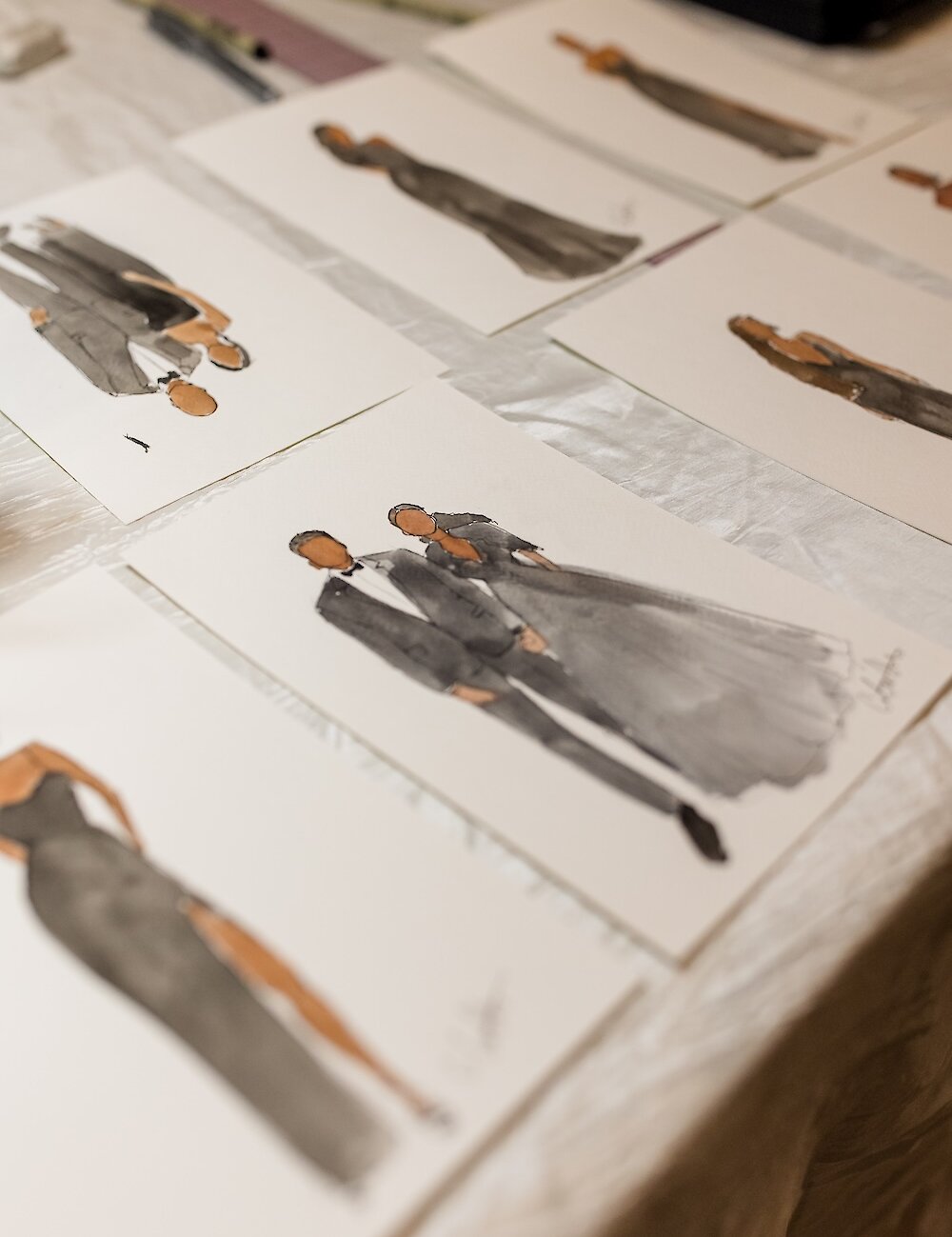 Several watercolor fashion illustrations of wedding guests, created by a live event artist, are laid out on a table as unique favors.