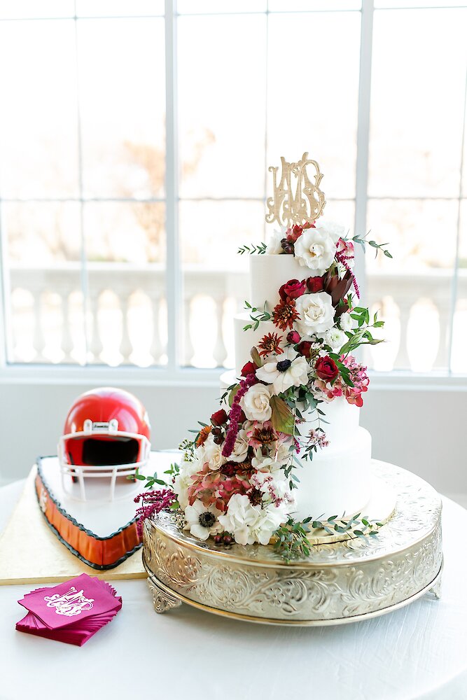 Elegant white tiered wedding cake with cascading red and white flowers beside a football helmet groom’s cake on a table.