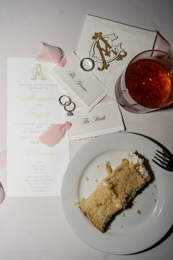 Flatlay of wedding invitation, bride and groom place cards, wedding rings, cocktail, and slice of wedding cake on a plate.