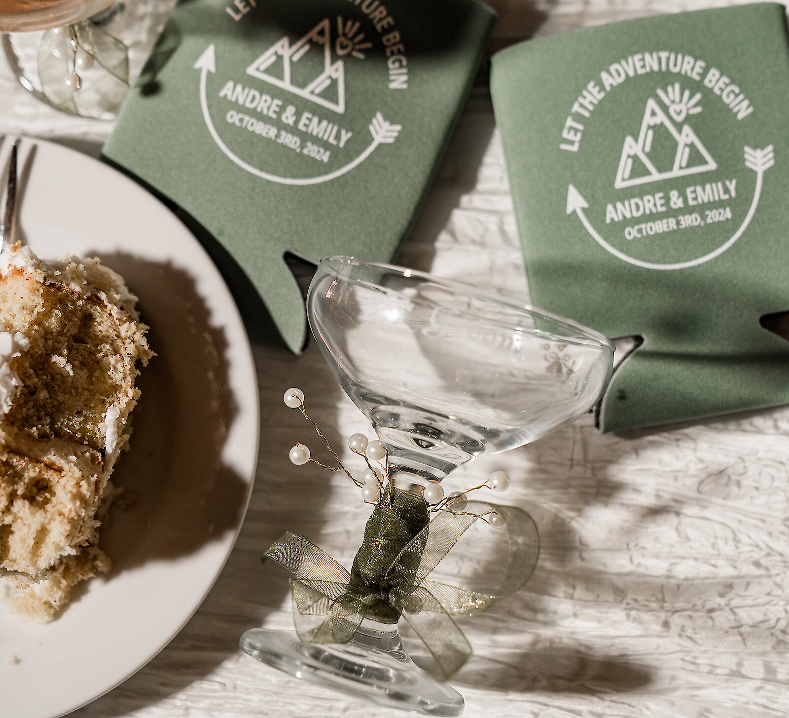 Wedding dessert table details with slice of cake on a plate, personalized green koozies reading “Let the Adventure Begin – Andre & Emily – October 3rd, 2024,” and a decorated glass with pearl accents