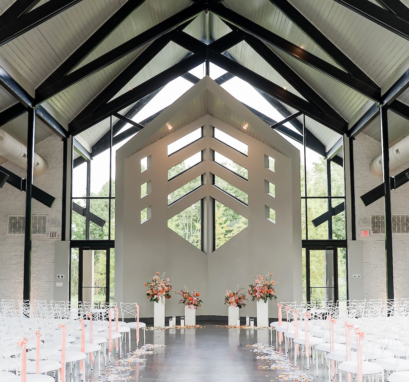 The modern interior of a wedding chapel with a high ceiling, exposed black beams, a unique altar structure, and rows of white chairs with peach ribbons.