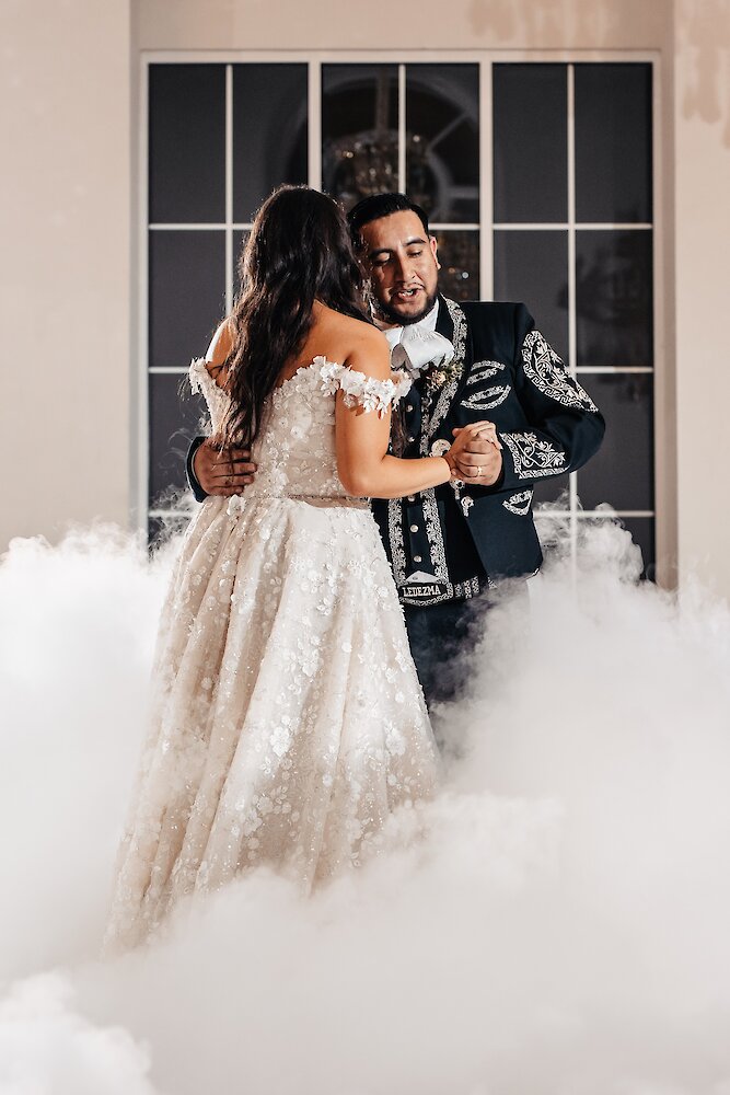 Bride and groom sharing their first dance surrounded by low-lying clouds, with the groom wearing a traditional mariachi suit and the bride in a floral off-the-shoulder wedding gown.