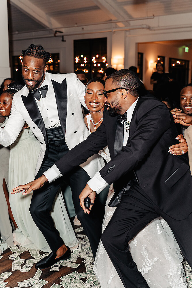 High-energy wedding reception dance photo with the bride, groom, and guests celebrating on a dance floor covered in scattered cash (money spray). The groom wears a white tuxedo jacket, and the bride is smiling amidst the excitement.