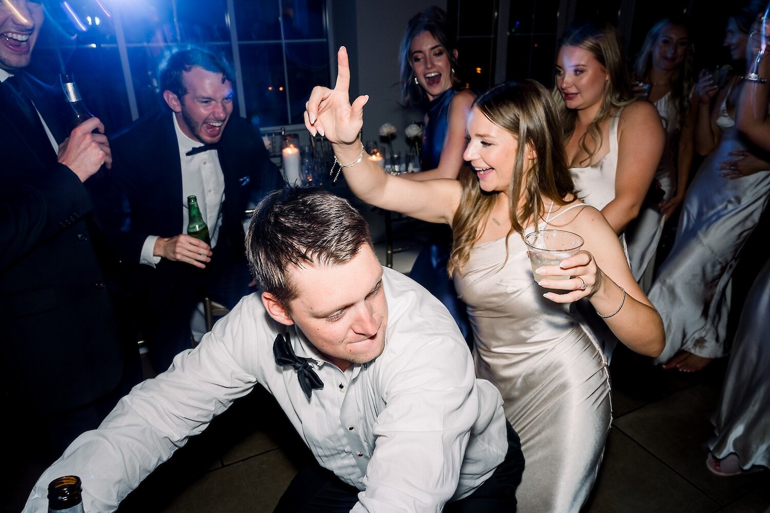 Lively wedding reception dance floor photo with guests cheering and dancing under blue and purple party lights. A bridesmaid in a champagne satin dress holds a drink, celebrating with a guest who is bending low.