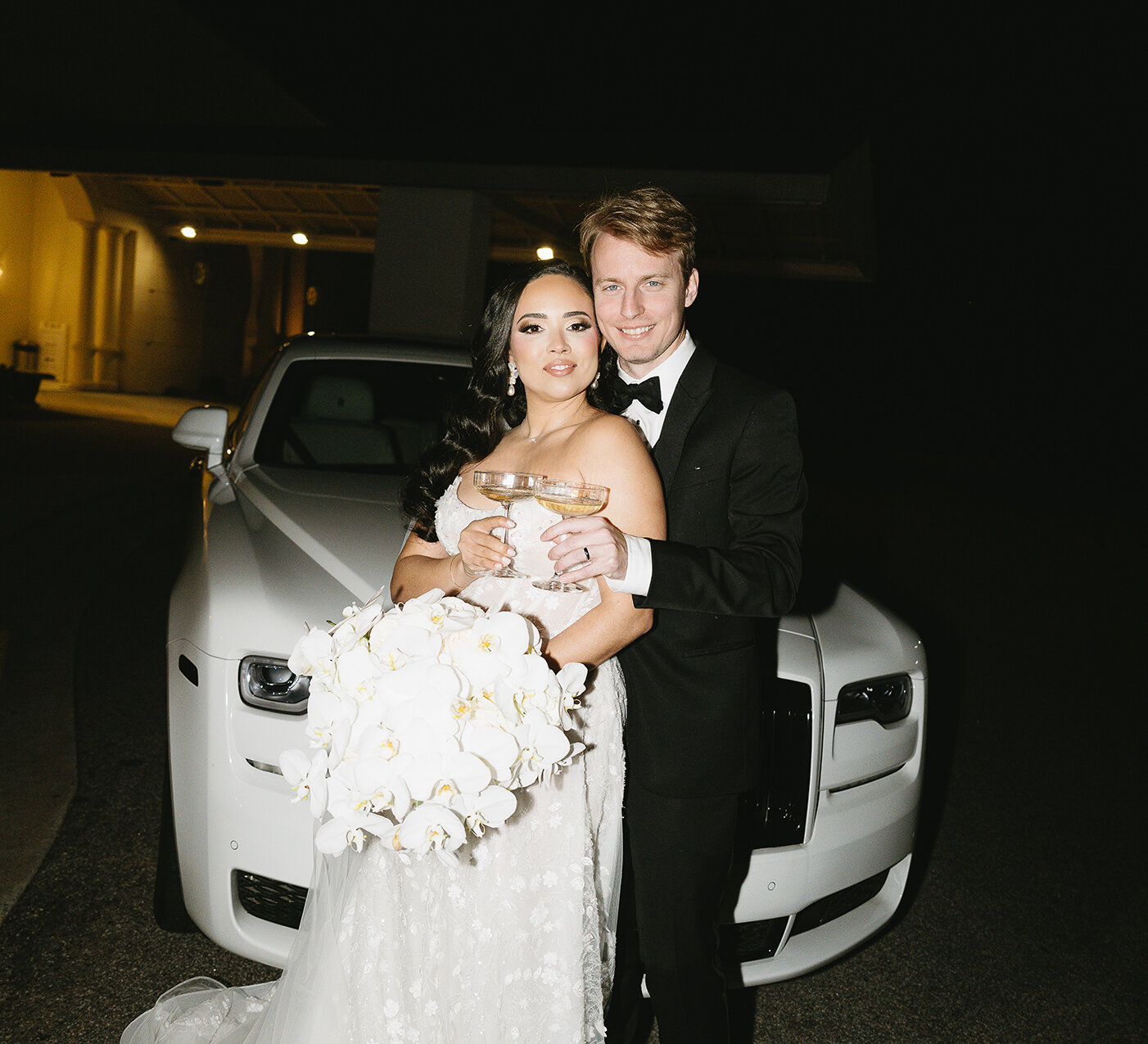Elegant nighttime wedding portrait of the couple toasting with champagne flutes in front of a white luxury car. The bride wears a strapless gown with floral appliqués and holds a massive white orchid bouquet. The groom wears a black tuxedo.