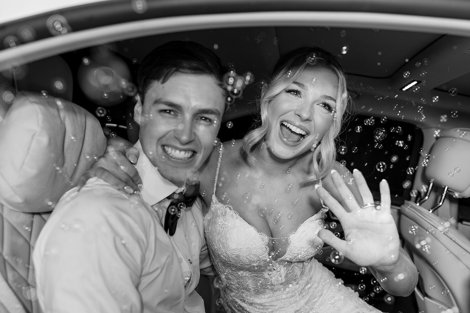 Joyful black and white close-up photo of the newlyweds smiling and waving from inside a white car, surrounded by exit bubbles. The bride wears a sequined lace gown and shows her wedding ring.