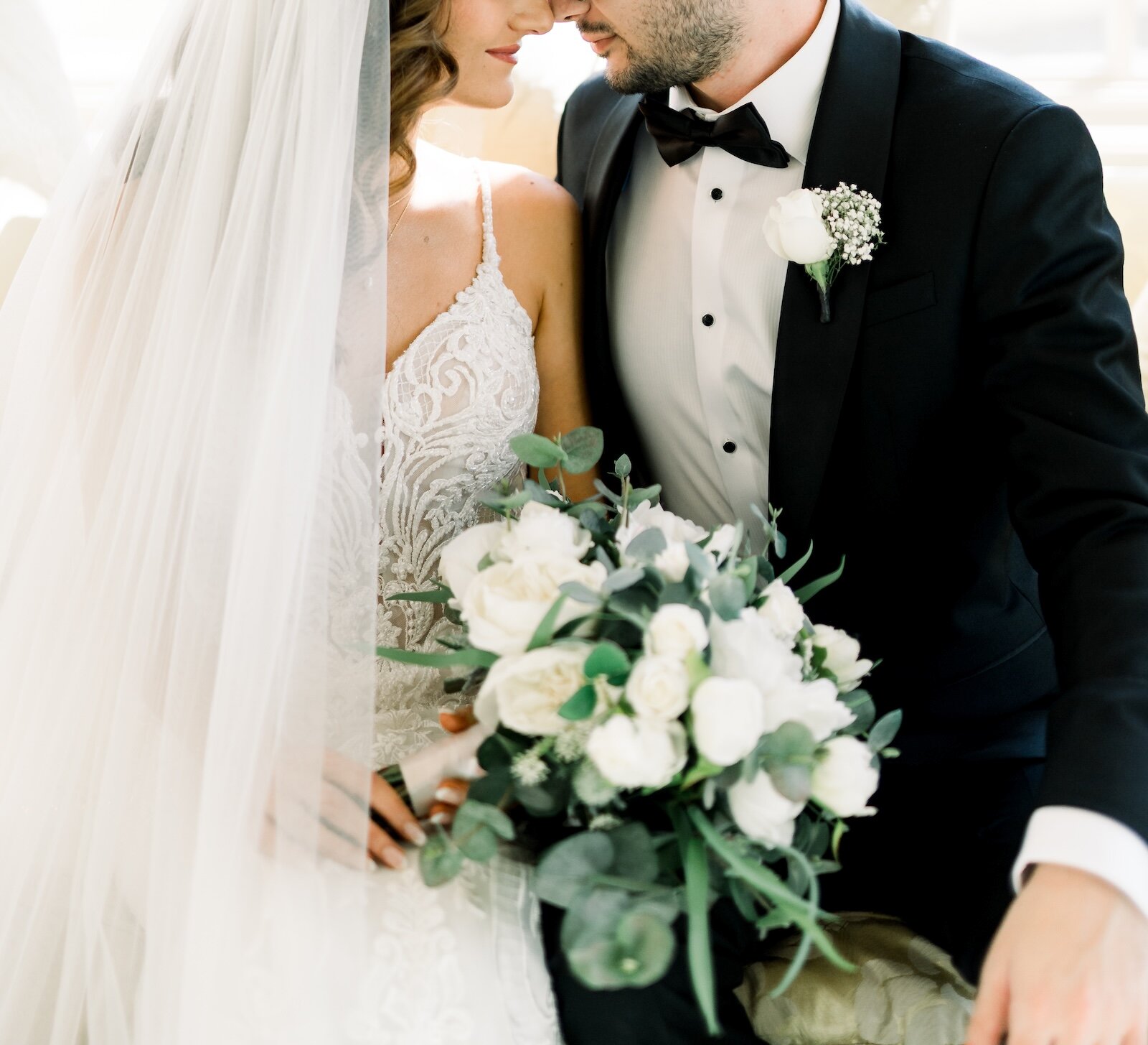 A close-up of a bride and groom embracing, holding a lush white rose and eucalyptus wedding bouquet.