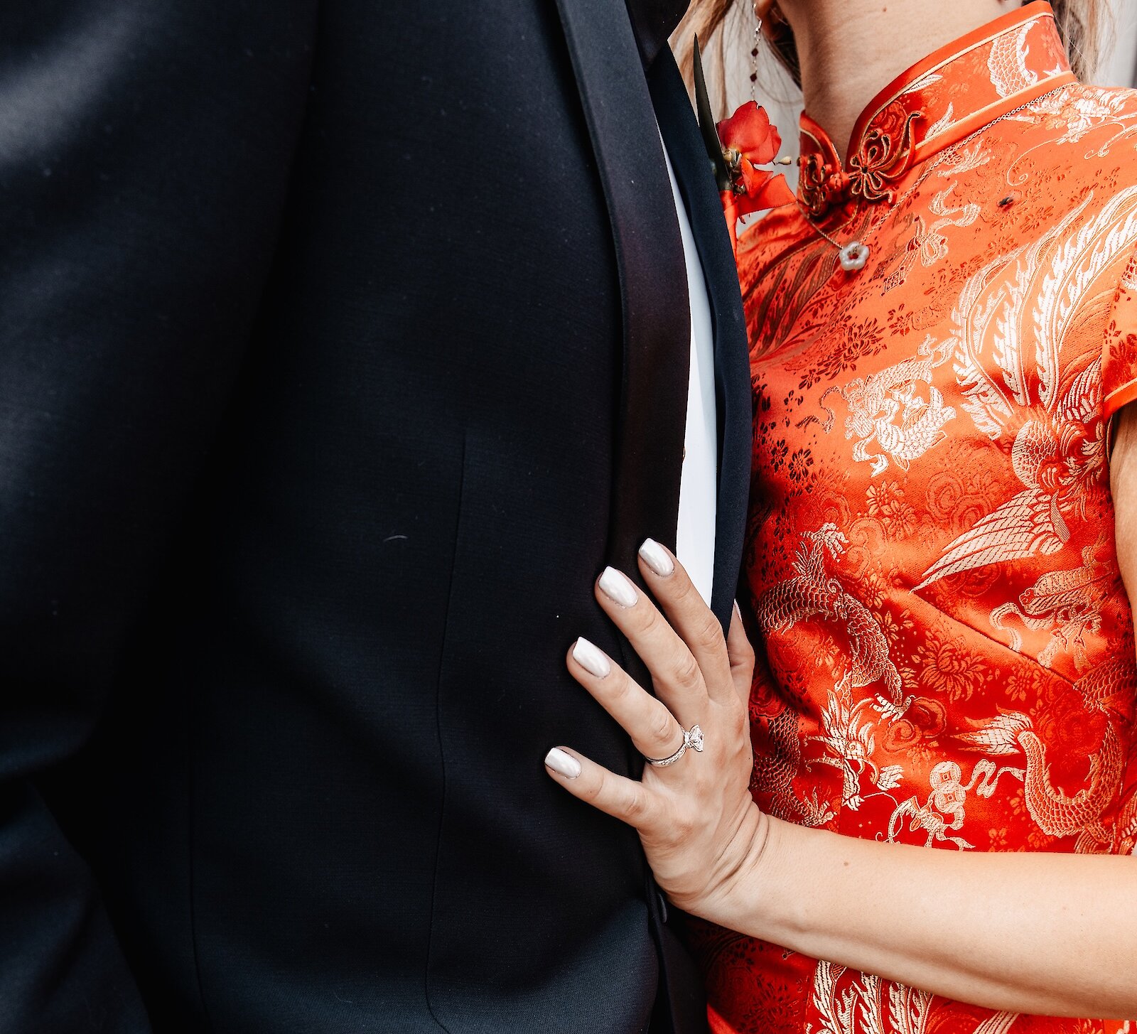 A close-up of a bride wearing a traditional red Chinese qipao with gold embroidery, resting her hand with an engagement ring on the groom's black suit.
