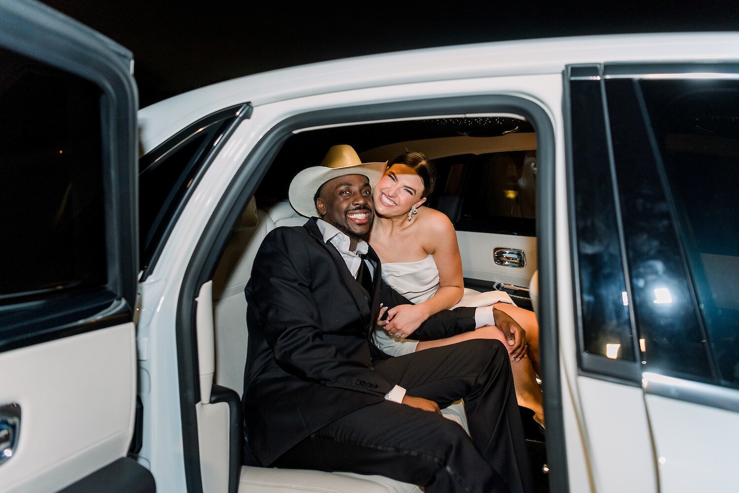Joyful nighttime photo of the newlywed couple smiling and embracing in the back seat of a white luxury car during their exit. The groom wears a tan cowboy hat and a black suit, and the bride wears a strapless satin gown.