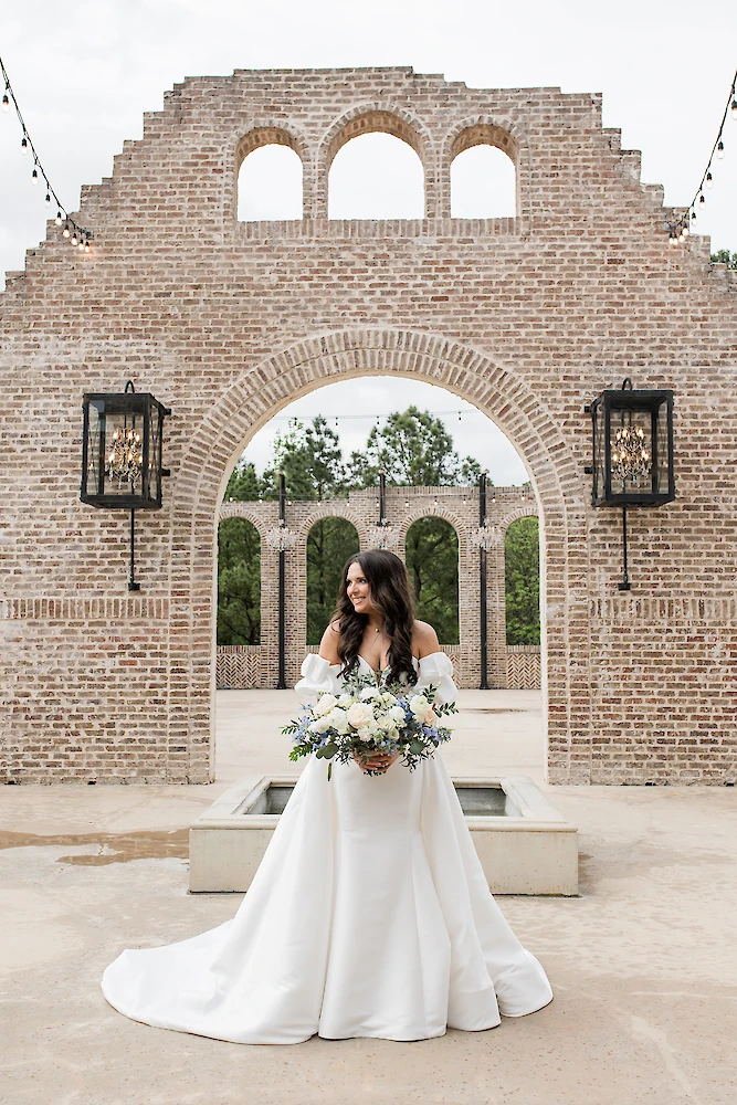 Stunning bridal portrait of the bride posing in an outdoor ceremony space featuring a towering, partially ruined brick wall with arched openings. The bride wears a voluminous off-the-shoulder satin gown and holds a large bouquet of white and blue flowers.