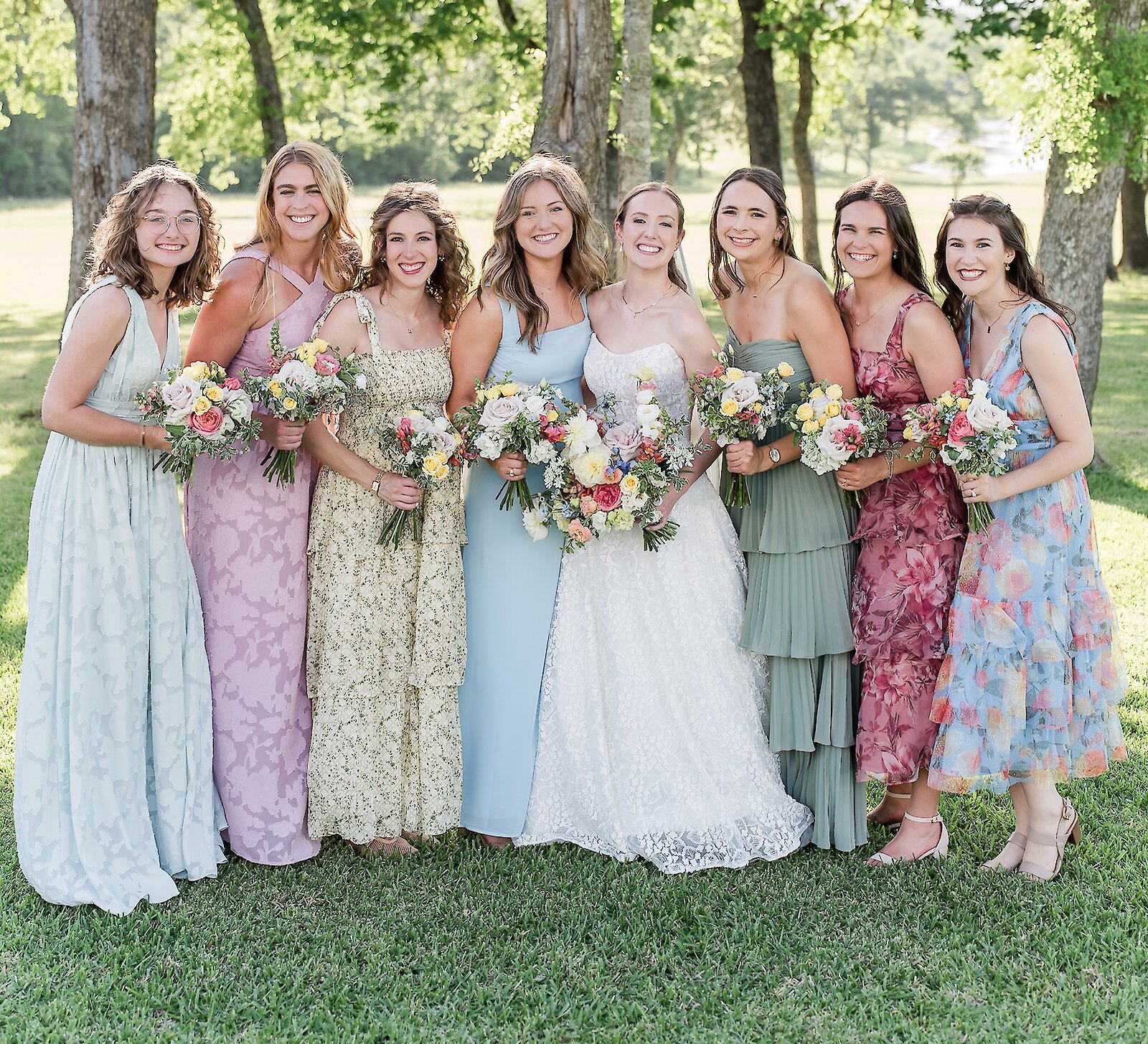 The bride poses with her seven bridesmaids, all wearing different pastel and floral patterned dresses, holding colorful bouquets in a grassy field.