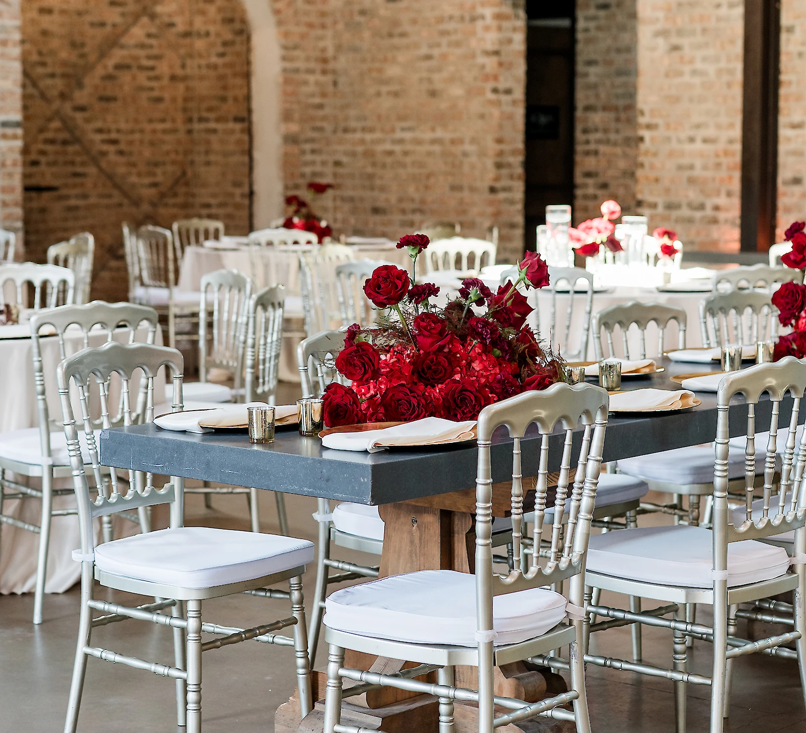 Elegant wedding reception table setting featuring a dark stone farm table surrounded by silver Chiavari chairs with white cushions. The centerpiece is a striking, lush arrangement of deep red roses, set against an exposed brick wall and arched entryway.