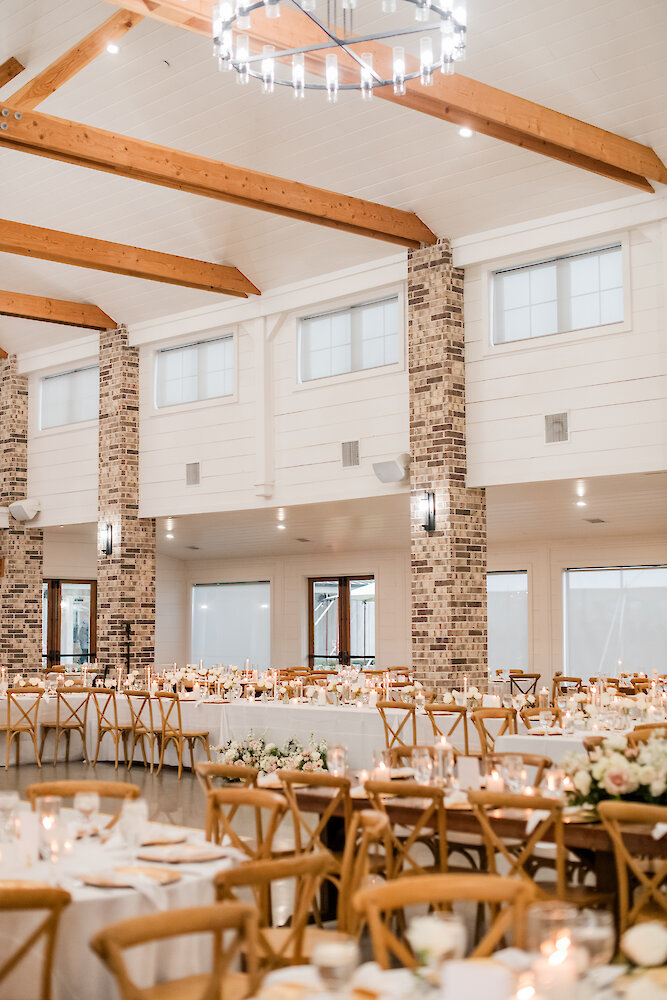 Warm and rustic elegant wedding reception hall featuring high white ceilings with exposed wooden beams and structural columns clad in light-colored brick. Tables are set with white linens, natural wood cross-back chairs, and candlelight for a cozy ambiance.
