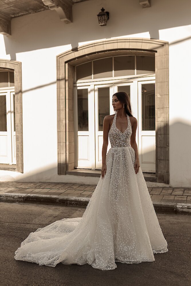 A beautiful bride models a white A-line wedding dress with a beaded halter neck bodice and a sweeping skirt outside a white building.