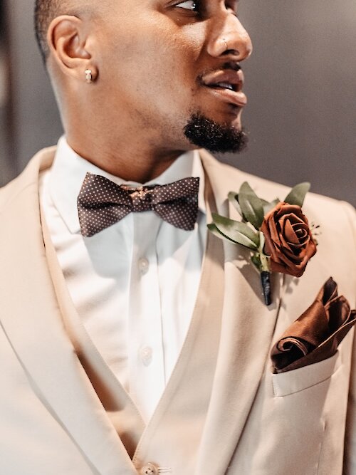 A close-up of the groom wearing a tan suit, a polka dot brown bow tie, and a dark rust-colored rose boutonniere.