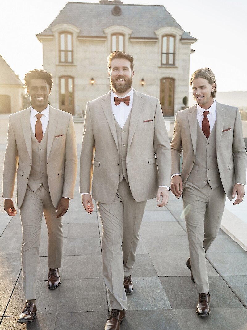 Three smiling groomsmen walk on a patio wearing light beige three-piece suits with rust-colored ties and bowties in front of a European-style building.