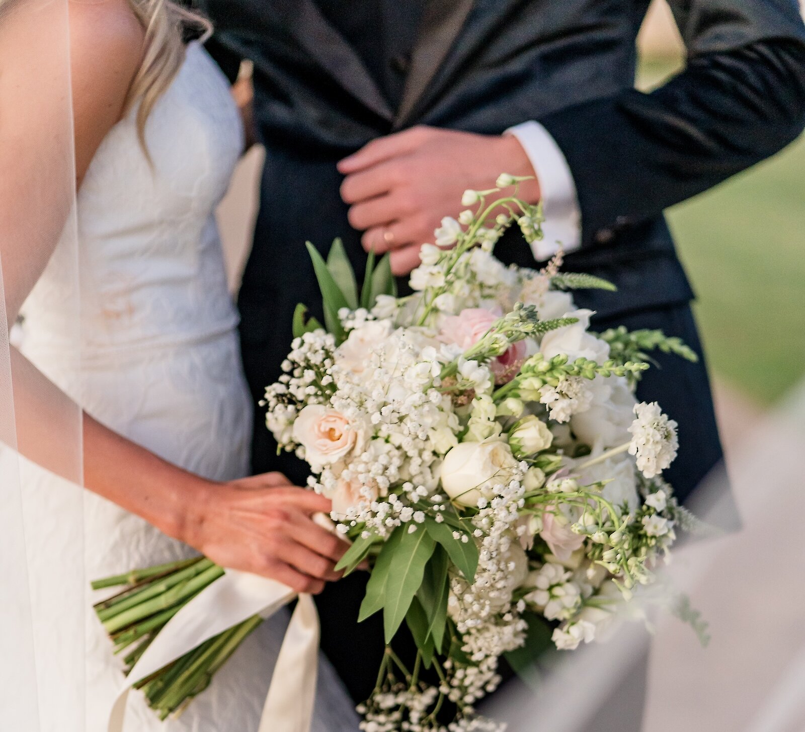 A bridal couple holding a white and pale pink bouquet with baby's breath and a light ribbon in a close-up shot.