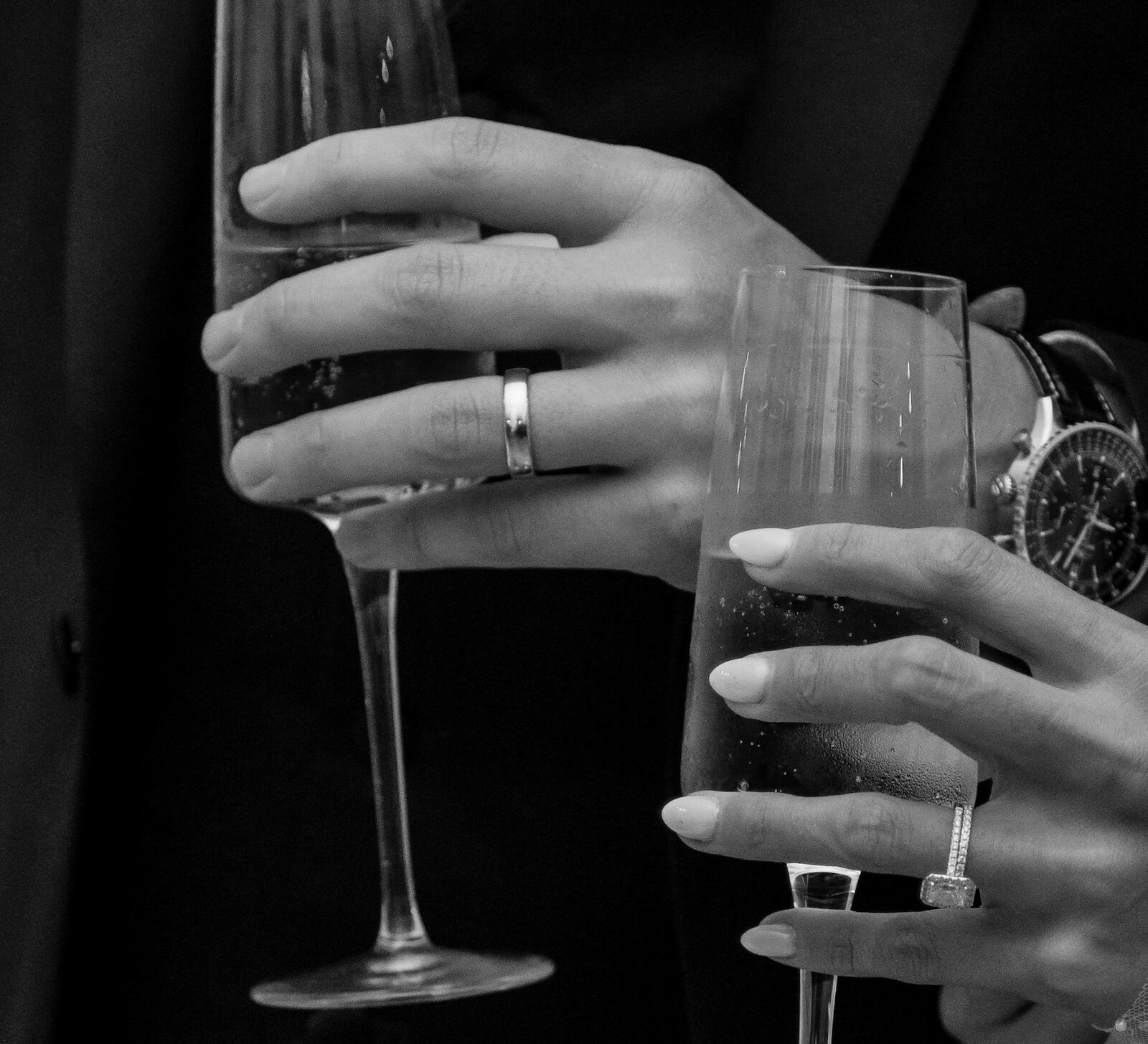 A black and white close-up of a couple's hands holding champagne flutes, showcasing their wedding and engagement rings.