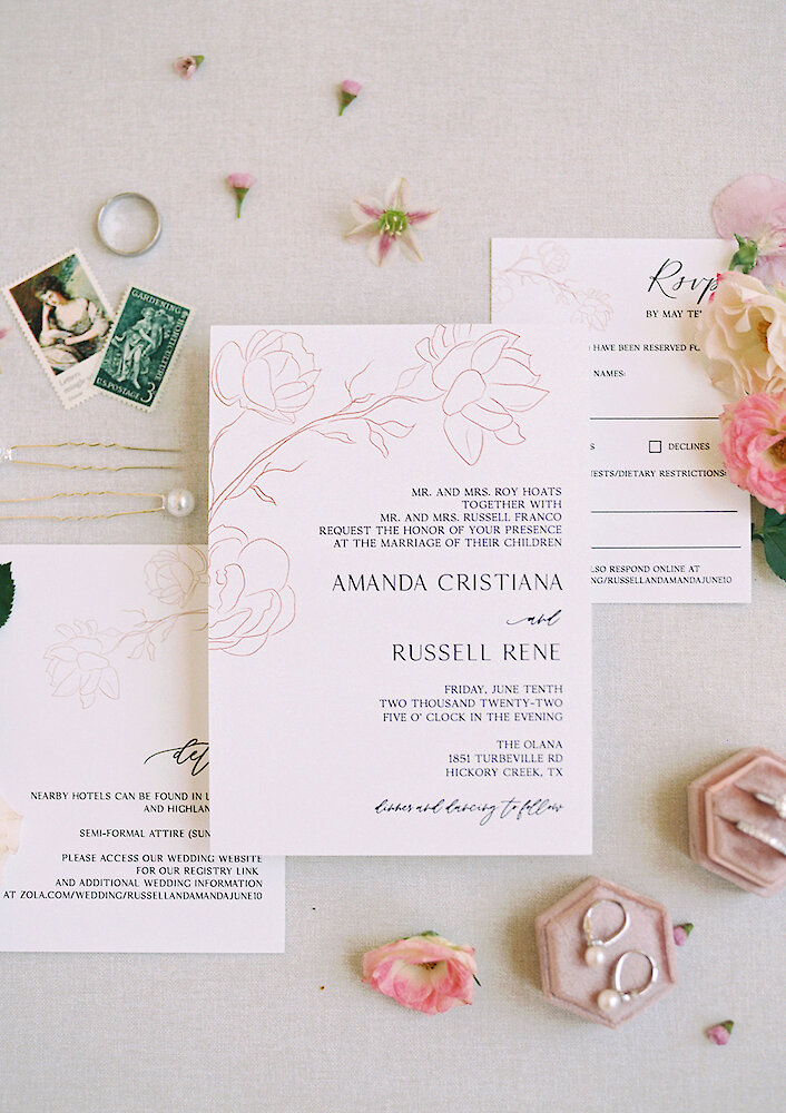 Close-up wedding tablescape detail featuring a gold-rimmed charger plate, gold flatware, and a blush pink velvet napkin holding a menu fan with a green tassel. The setting is framed by a lush, colorful centerpiece of pink, peach, and coral roses.
