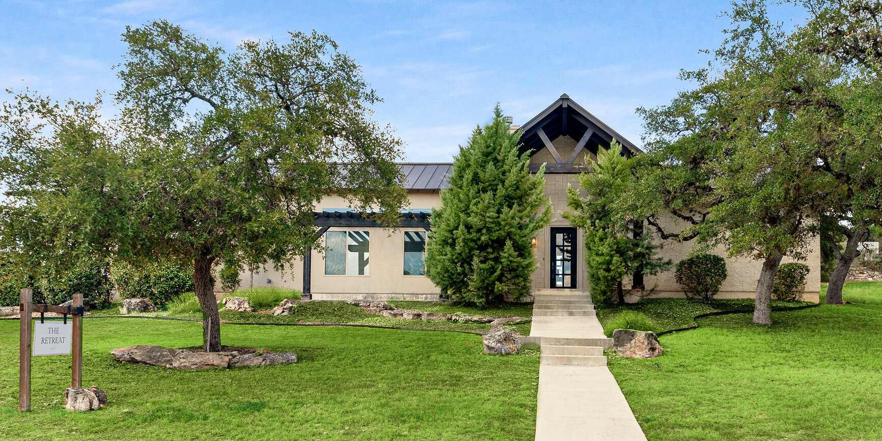 Exterior view of a modern retreat building with stone pathway, large trees, and landscaped green lawn under a clear sky.