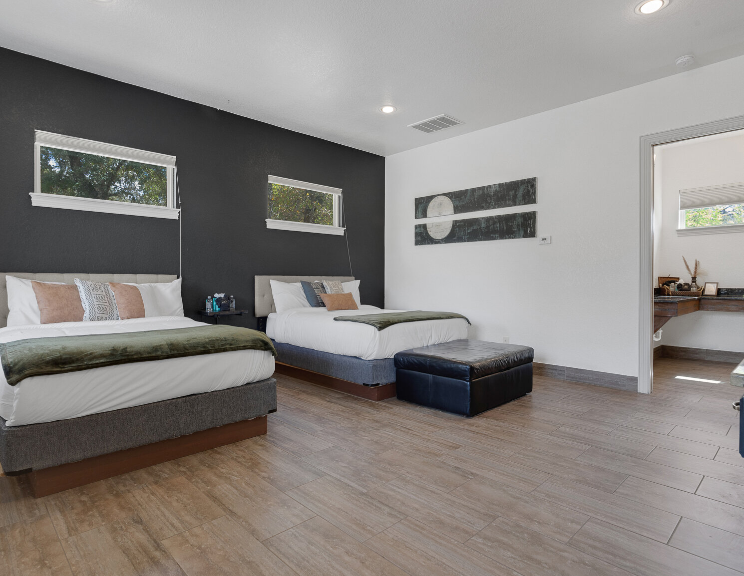 Modern bedroom with two queen beds, dark accent wall, wood-look tile flooring, and adjoining bathroom.