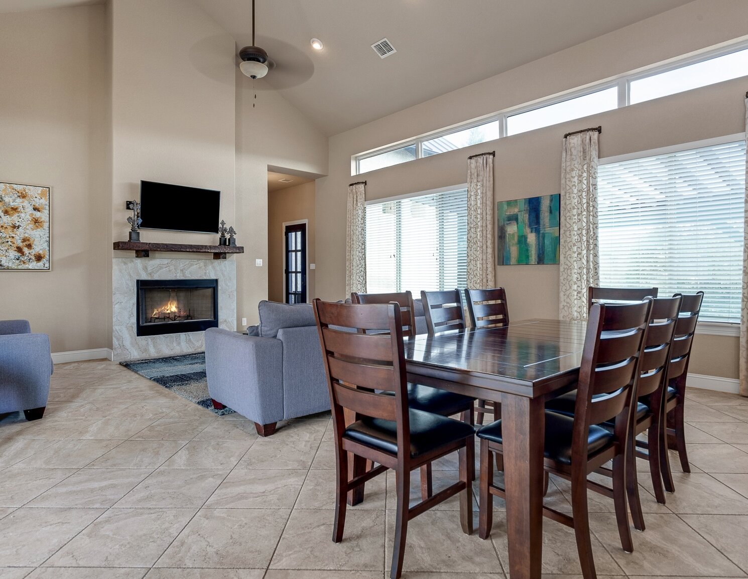A combined living and dining room with tiled floors, featuring a dark wood dining table for eight and a living area with a stone fireplace and television.