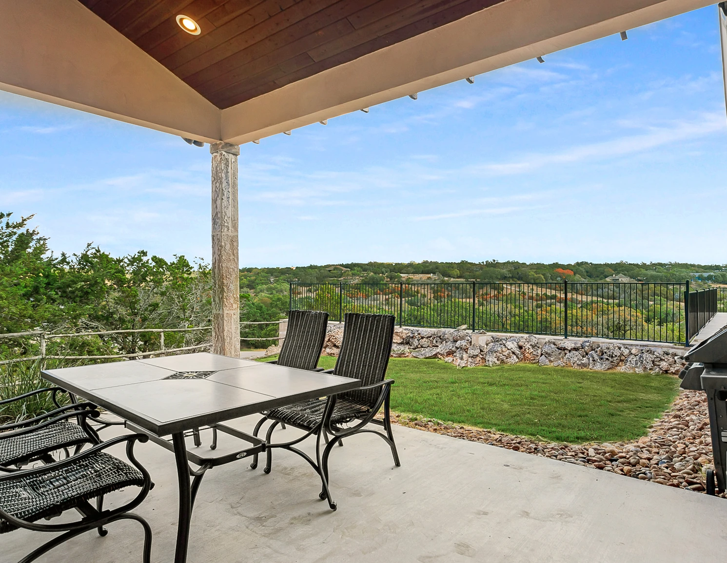 Covered patio with outdoor dining set, grill, and scenic hill country view behind a metal fence.