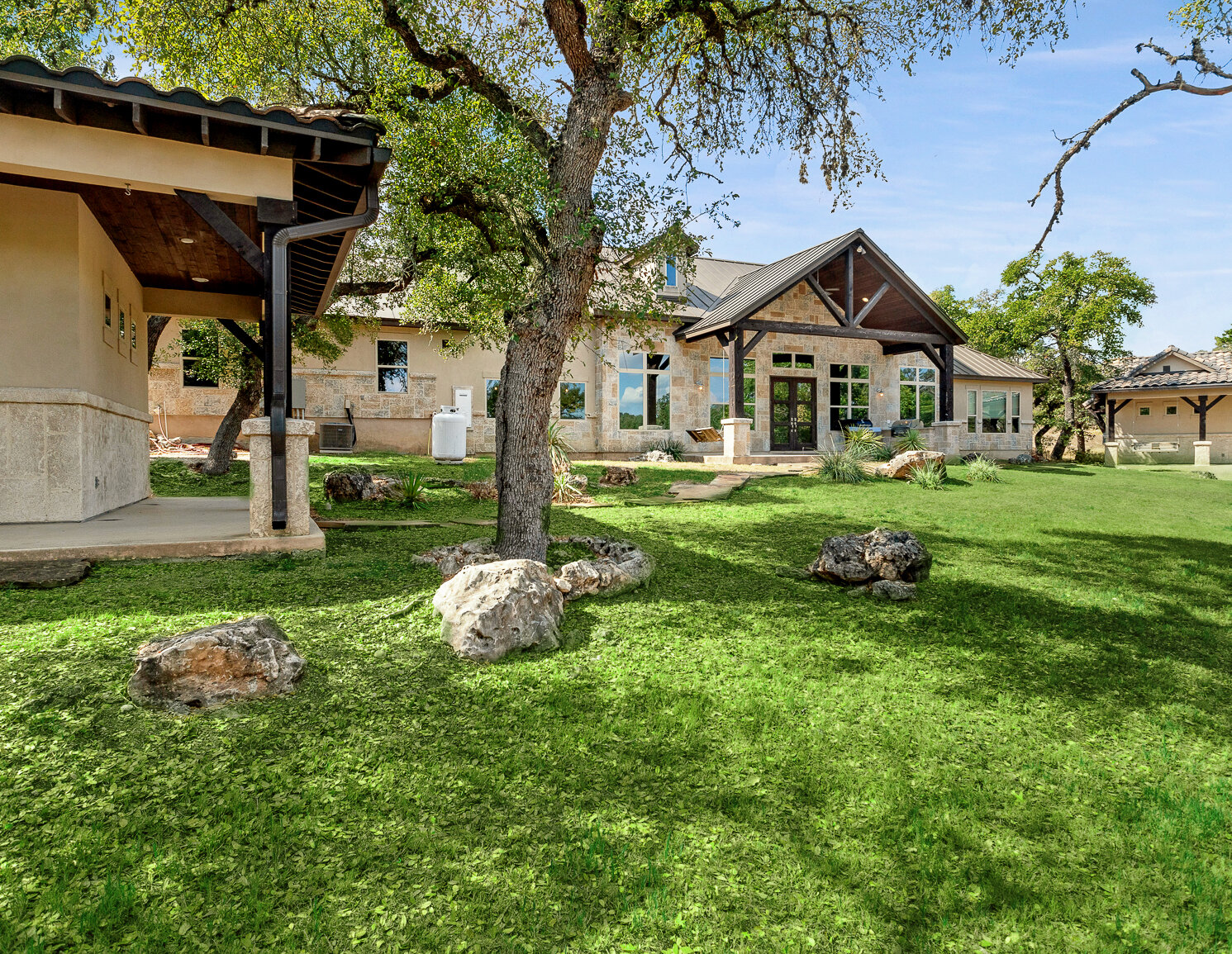 Spacious hill country estate with stone exterior, large windows, covered patios, and mature oak trees on a green landscaped lawn.