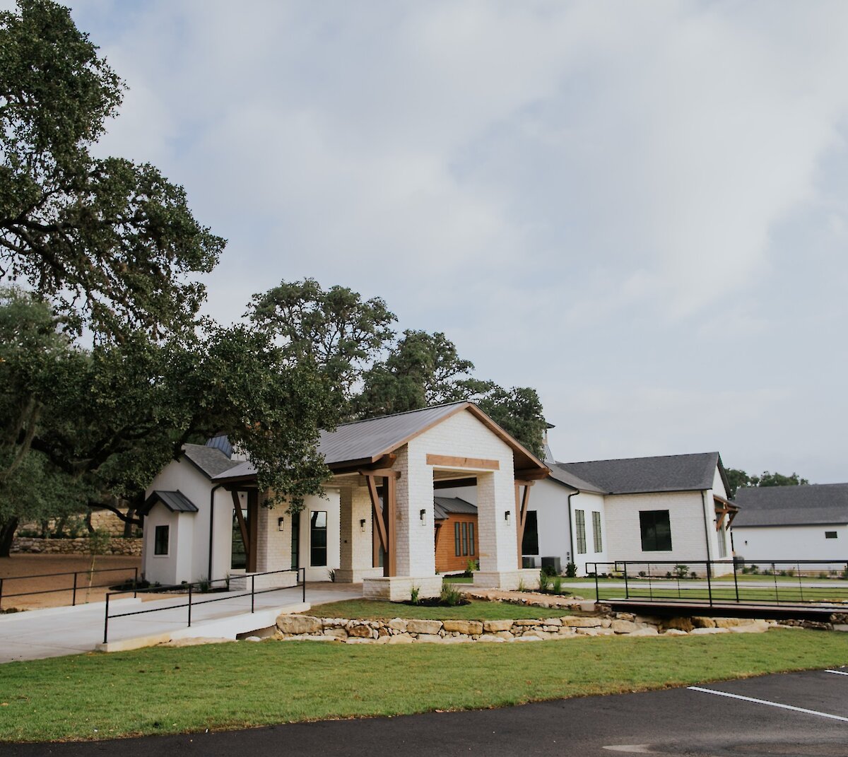 Modern white ranch-style venue building with wood accents, surrounded by oak trees and landscaped stone features