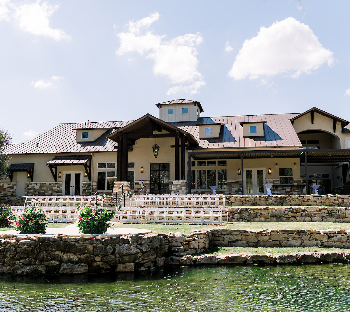 Outdoor view of a rustic stone and stucco wedding venue with tiered landscaping, water feature, and ceremony chairs set up on the lawn.