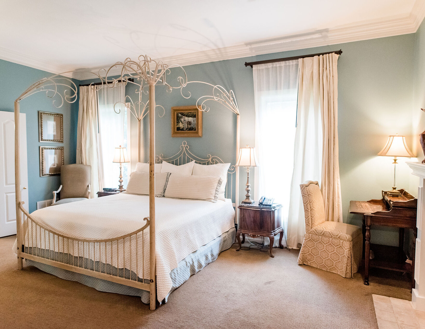 Bright and elegant bedroom featuring a decorative four-poster metal bed, soft blue walls, white bedding, vintage wooden furniture, and natural light from tall curtained windows.