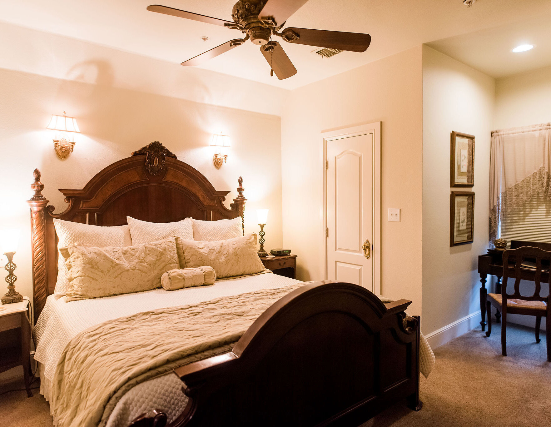 Warm, elegant bedroom with a carved wooden bed frame, neutral bedding, bedside lamps, and a small writing nook with a vintage desk and chair.