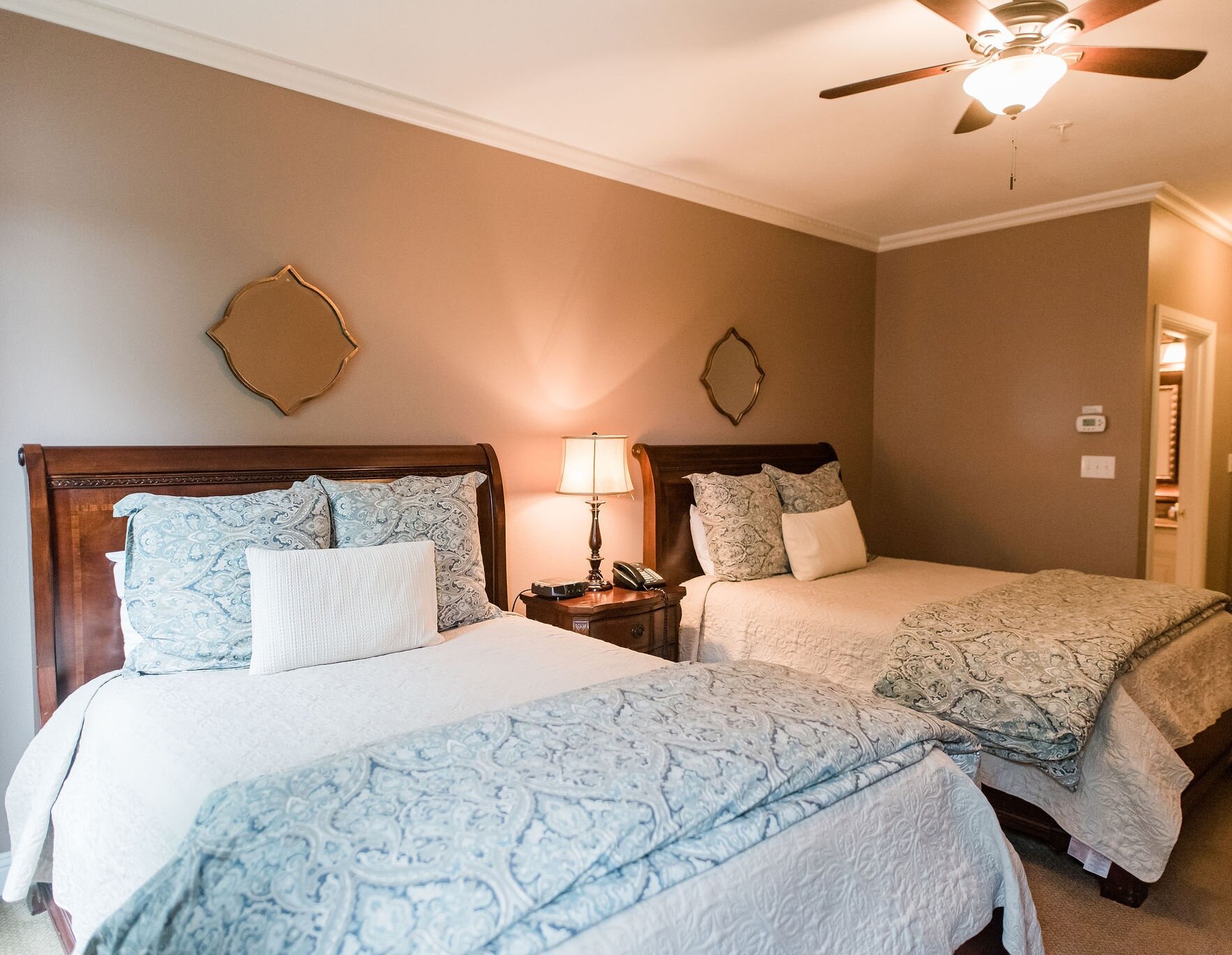 A hotel-style guest room featuring two wooden beds with blue and white bedding, a nightstand, and two decorative mirrors on a tan wall.