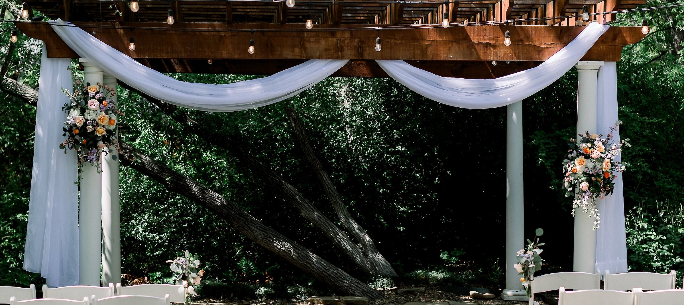 An outdoor wedding ceremony space featuring white folding chairs lining a flagstone aisle leading to a wooden pergola draped with white fabric and string lights.