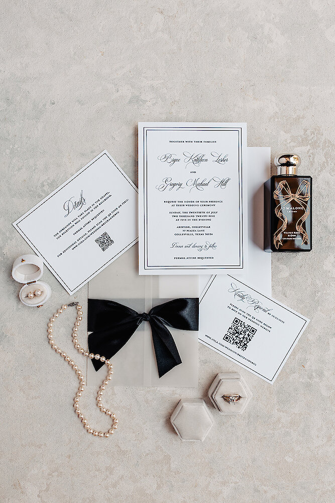 Flat lay of a classic wedding invitation suite with black and white typography, resting on a textured light background. Details include a black ribbon, pearl necklace, pearl earrings, wedding rings in velvet boxes, and a Jo Malone perfume bottle.