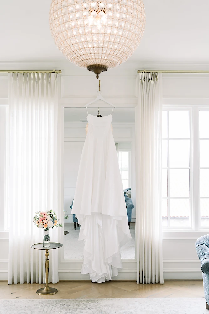 Elegant bridal details photo showing a simple strapless white satin wedding gown hanging on a large mirror in a bright suite with white window treatments. A blush and peach floral bouquet sits on a gold side table, beneath a shimmering crystal chandelier.