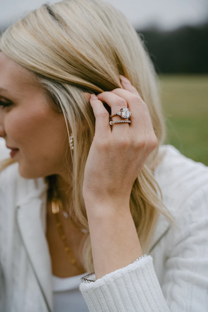 Close-up photo of an engagement ring featuring a large, horizontal emerald-cut diamond with side stones, stacked with a diamond wedding band, worn on the hand of a blonde woman wearing a white knit sweater.