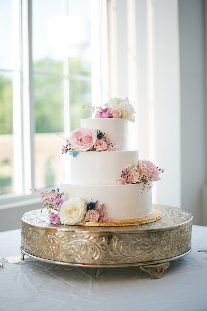 Three-tiered white wedding cake with smooth buttercream frosting, decorated with fresh pink, white, and lavender roses and thistle accents, sitting on an ornate silver and gold mirrored cake stand in front of a window.