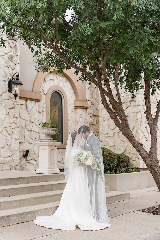 Intimate wedding portrait of the bride and groom kissing beneath her sheer veil outside a Mediterranean-style stone chapel. The groom wears a gray suit, and the bride holds a blush and white floral bouquet, standing on a sidewalk near stone steps.