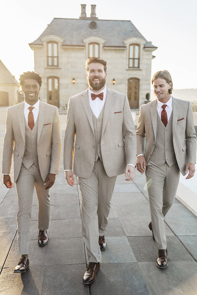 Stylish portrait of three men walking toward the camera on a rooftop terrace during sunset. They are wearing matching light beige three-piece suits with burgundy ties and bowties, against the backdrop of a European-style stone estate.