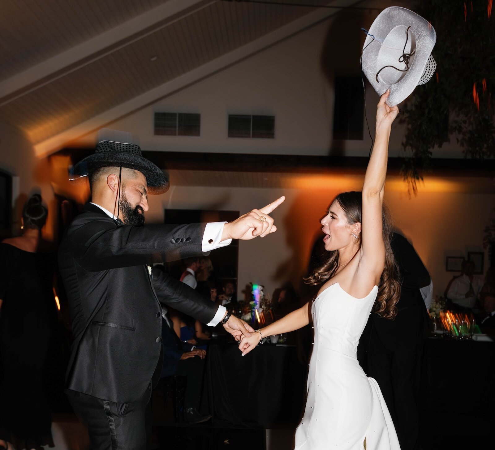 The bride and groom are enthusiastically dancing at their reception, with the groom wearing a black hat and the bride holding a white fedora hat raised above her head.