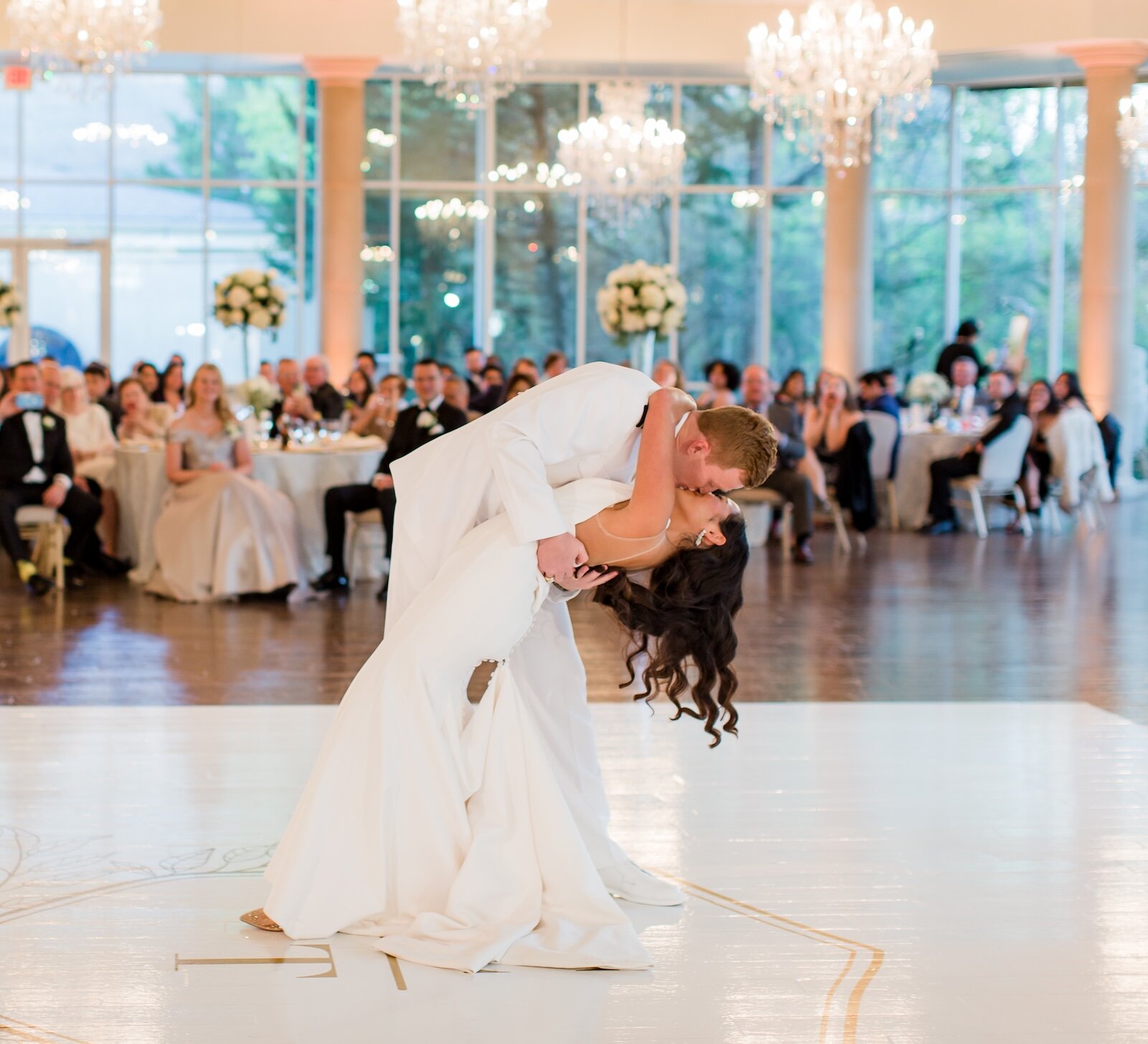 The bride and groom share a passionate kiss while dipping during their first dance on a custom dance floor in a grand ballroom with crystal chandeliers.