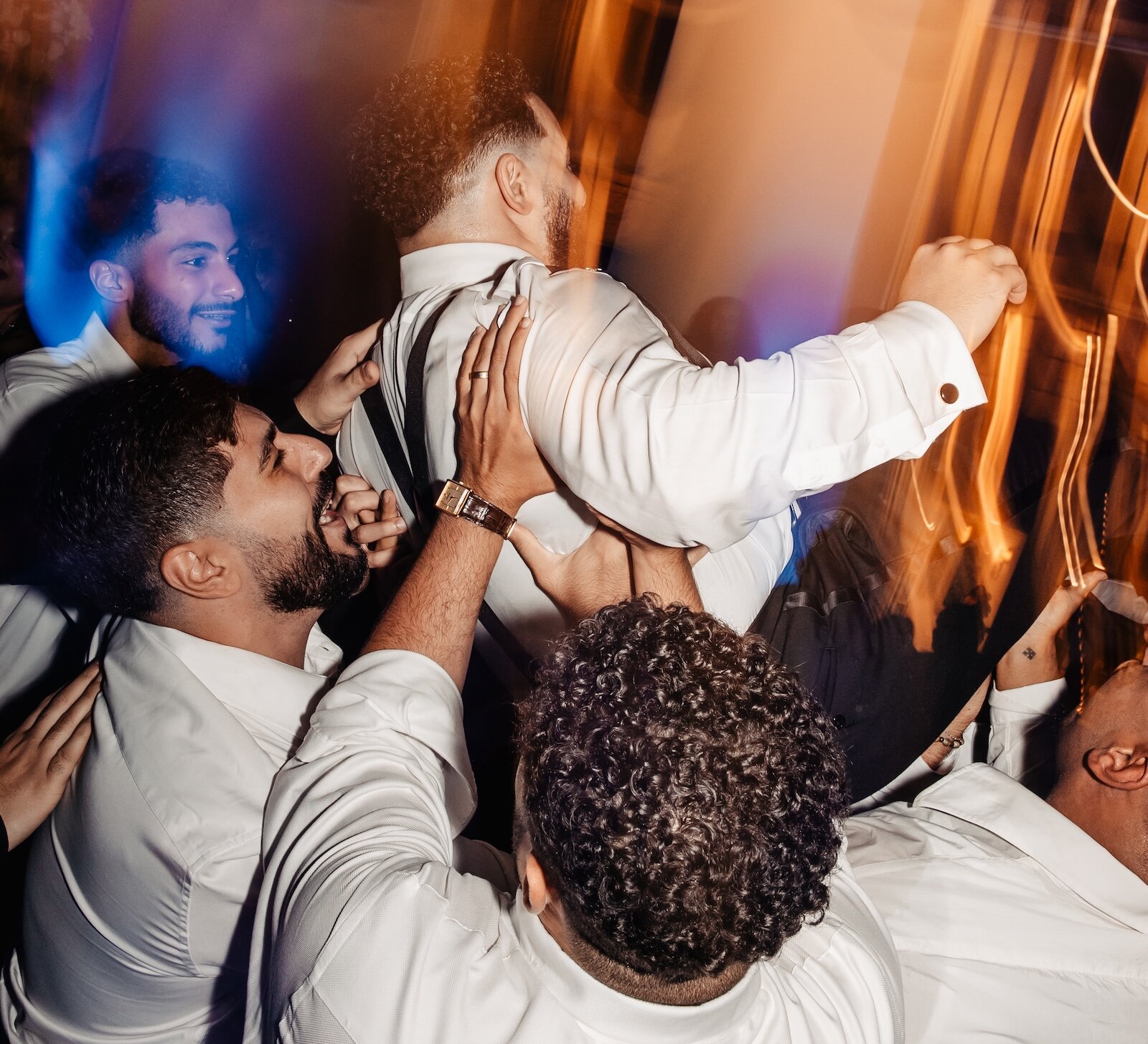 The groom is lifted into the air by his friends during the traditional Hora dance at the wedding reception, illuminated by dramatic orange and blue lighting.