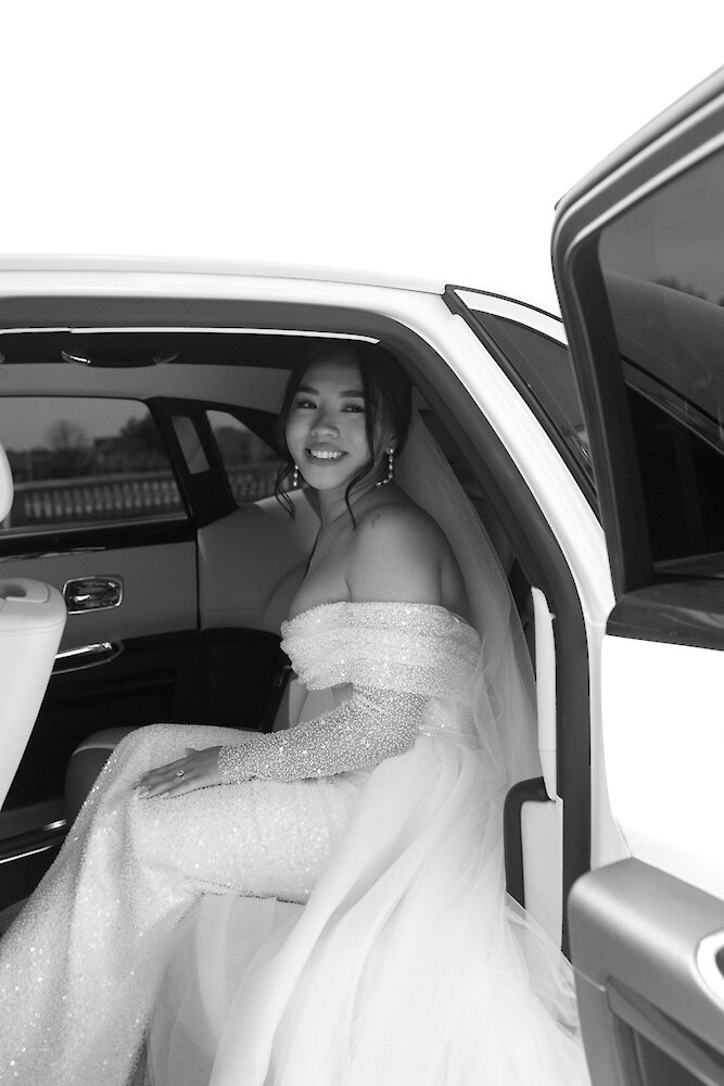 A smiling bride wearing a sparkling off-the-shoulder gown sits in the back seat of a white Rolls Royce getaway car in a black and white photograph.