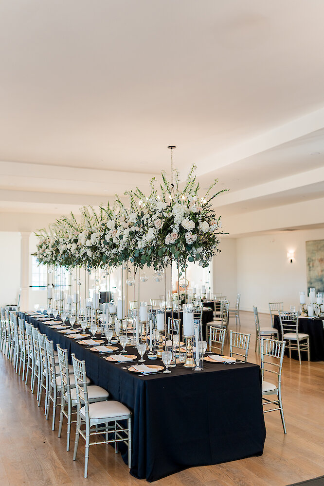 Elegant wedding head table set with black linens, gold charger plates, and silver Chiavari chairs. The table features a row of spectacular tall white and green floral arrangements suspended overhead, creating a dramatic focal point in the bright reception room.