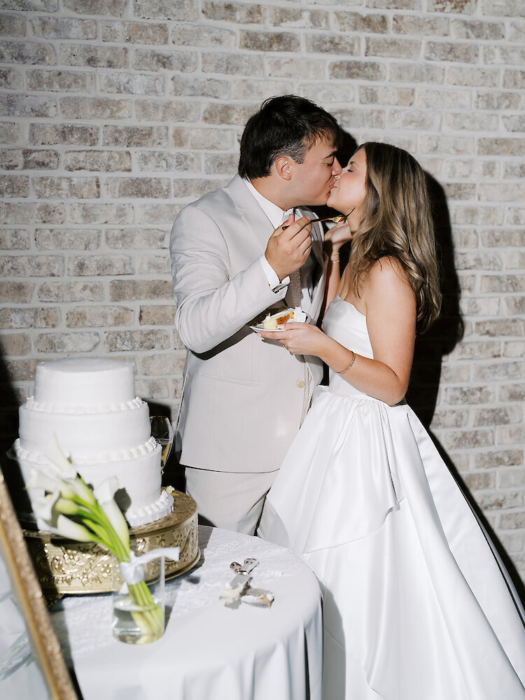 The bride and groom kiss while eating a bite of cake next to their white tiered wedding cake and a rustic brick wall.