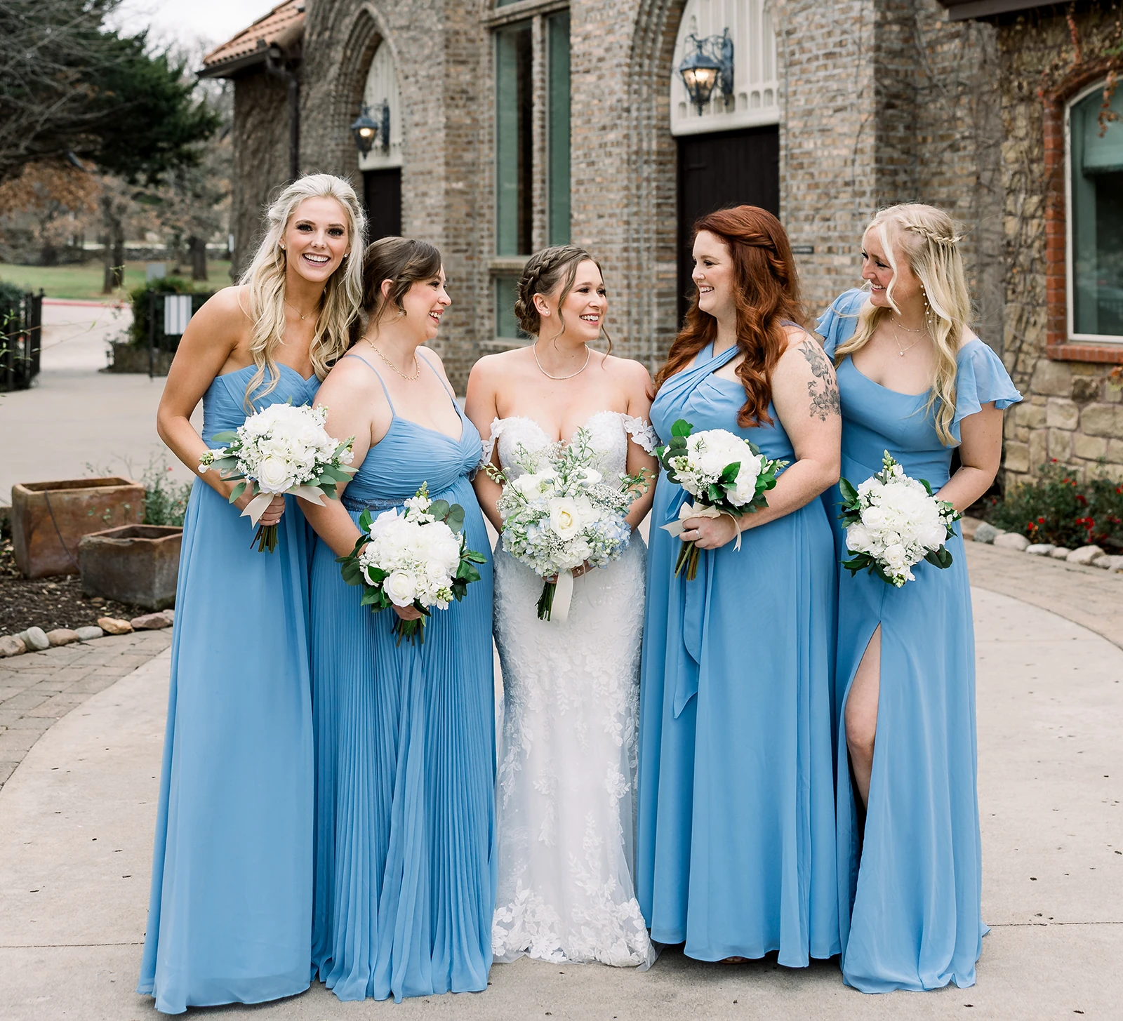 Bride smiling with her bridesmaids dressed in light blue gowns, holding white floral bouquets, standing outside a Mediterranean-style stone wedding venue