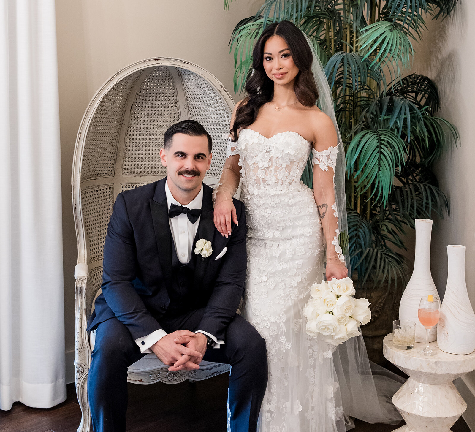 Bride and groom posing together indoors, with the groom seated and the bride standing beside him in a lace off-the-shoulder wedding gown holding a white rose bouquet